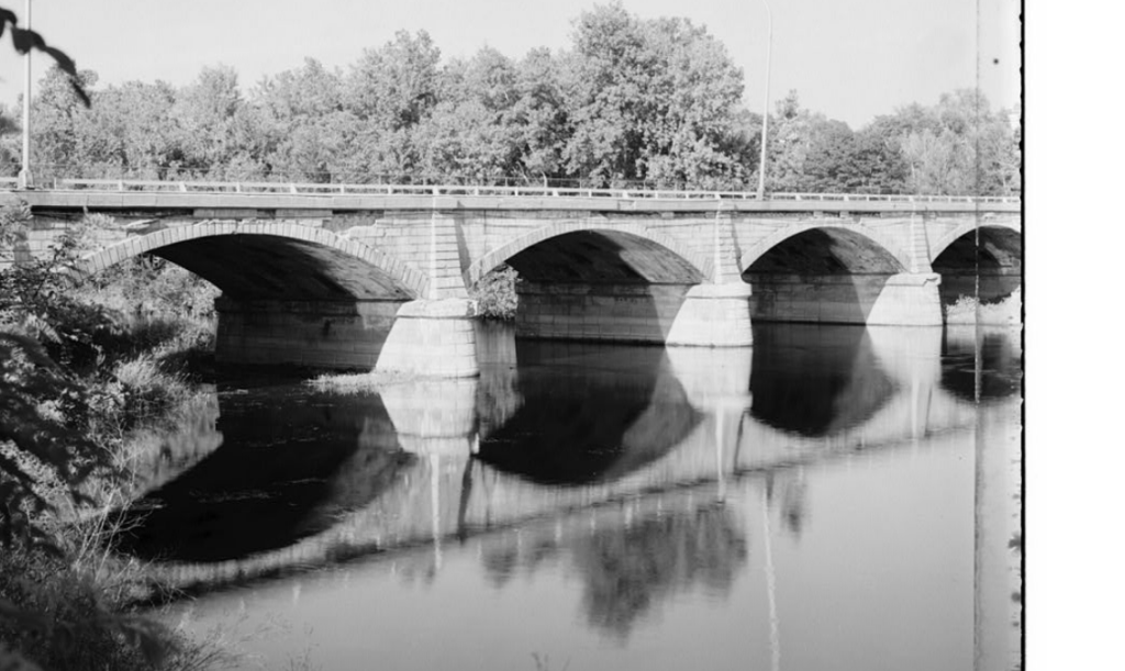 Black and white photo of four-arch stone bridge over river with trees in background. [Historic American Engineering Record, C., Kasson, M. O., Burr, W. H. & Melan, J. (1968) Sandy Hill Bridge, Bridge Street, spanning Hudson River, Hudson Falls, Washington County, NY. Hudson Falls New York Washington County, 1968. Documentation Compiled After. [Photograph] Retrieved from the Library of Congress, https://www.loc.gov/item/ny1549/.]